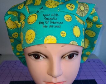 May include: A teal scrub cap featuring a repeating pattern of yellow suns with various facial expressions. The front band displays the black text, Your little Sarcastic Ray Of Sunshine Has Arrived!, with a decorative zigzag stitch line below it.