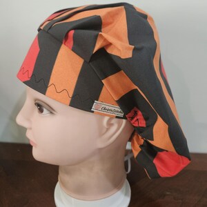 May include: A patterned scrub cap in orange, red, and black, worn by a mannequin head. The cap features a geometric design and the text "ChristieStitches" on a small tag.