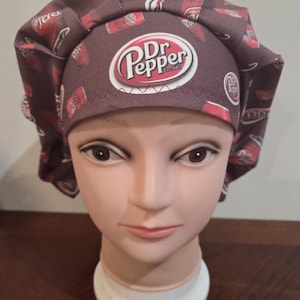 May include: A burgundy scrub cap featuring a repeating pattern of Dr Pepper cans and logos. The cap has a prominent Dr Pepper logo on the front, with a white oval and red accents. The cap is designed for medical professionals or those in the healthcare field.