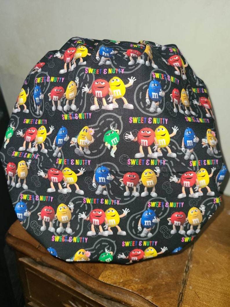 Pu&ograve; includere: Tessuto nero con un motivo ripetuto di personaggi M&M's colorati. I personaggi indossano scarpe e si tengono per mano. Il tessuto ha la scritta "Sweet & Nutty" stampata.