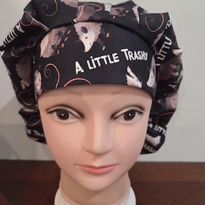 May include: A black scrub cap featuring a repeating pattern of opossums and the white text A LITTLE TRASHY printed on the front band. The fabric hat has a pleated design around the crown and is displayed on a mannequin head for clear visibility.