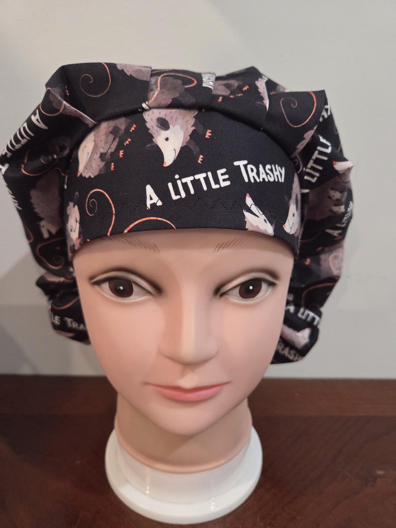 May include: A black scrub cap featuring a repeating pattern of opossums and the white text A LITTLE TRASHY printed on the front band. The fabric hat has a pleated design around the crown and is displayed on a mannequin head for clear visibility.