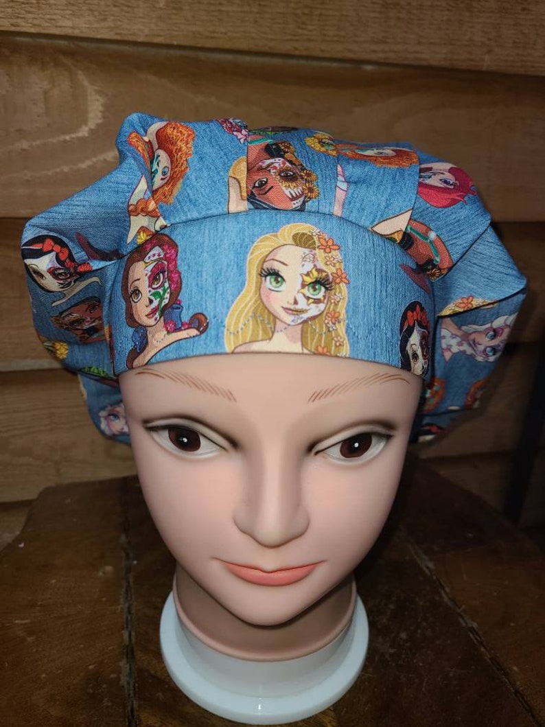 May include: A blue scrub hat with a print of Disney princesses with Day of the Dead makeup. The princesses include Rapunzel, Belle, and Ariel.