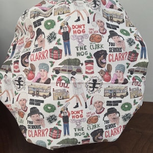 May include: A white scrub cap featuring a repeating pattern of Christmas-themed movie characters, station wagons, turkeys, and text including "Don't hog the nog," "Save the neck for me Clark," "Shitters full," and "You serious Clark?" with holly accents.