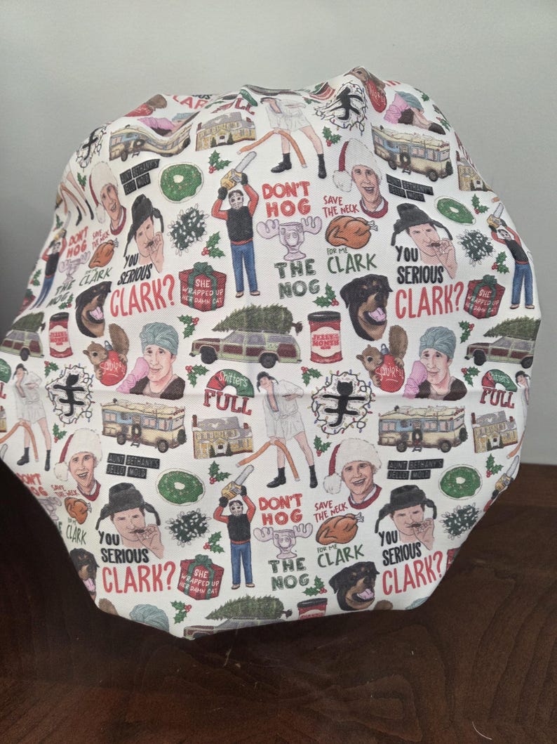 May include: A white scrub cap featuring a repeating pattern of Christmas-themed movie characters, station wagons, turkeys, and text including "Don't hog the nog," "Save the neck for me Clark," "Shitters full," and "You serious Clark?" with holly accents.
