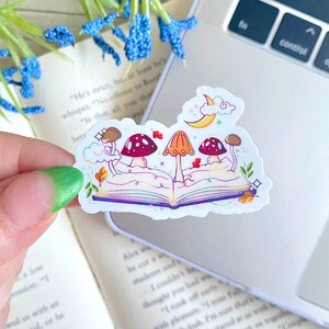 May include: A sticker featuring a whimsical illustration of an open book with three mushrooms growing from its pages. The book is surrounded by stars, clouds, and leaves. The mushrooms are red, orange, and brown. The sticker is perfect for adding a touch of magic to your planner, laptop, or water bottle.
