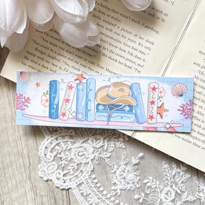 Peut inclure: Un marque-page fantaisiste représentant une étagère avec des livres illustrés, un chapeau de cow-boy et des coquillages. Le design incorpore des tons bleus, roses et beiges, créant une esthétique charmante. Pour les amateurs de livres.