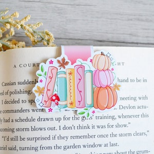May include: A pink and white bookmark with a stack of colorful books and pumpkins. The bookmark has a floral design and a glittery finish.