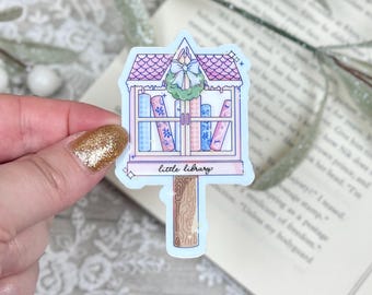 May include: A sticker featuring a whimsical illustration of a miniature library. The library has a pink roof, a green wreath, and shelves filled with colorful books. The text "little library" is at the bottom.