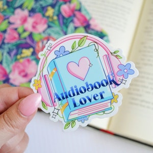 May include: A sticker with a blue book, pink headphones, and flowers. The book has a pink heart and the text "Audiobook Lover" in blue.