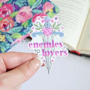 May include: A white sticker with a pink and purple gradient design. It features a stylized dagger with a pink heart and vines with flowers. The text "enemies to lovers" is written in a pink and purple gradient font.