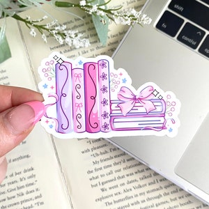 May include: A sticker featuring a stack of books and a row of books in shades of pink and purple, adorned with bows and floral accents. The sticker is held over a laptop and an open book.