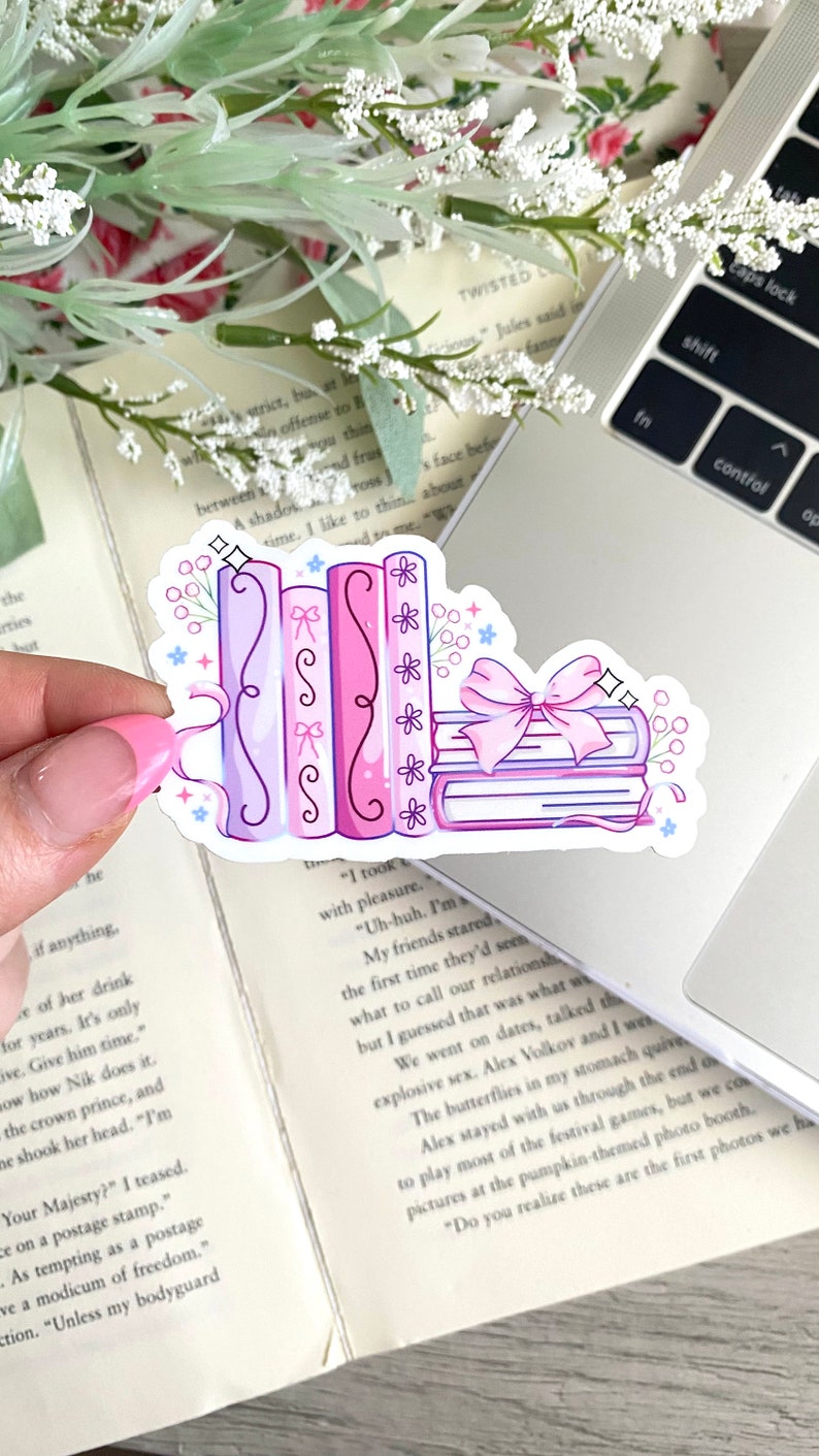 May include: A sticker featuring a stack of books and a row of books in shades of pink and purple, adorned with bows and floral accents. The sticker is held over a laptop and an open book.