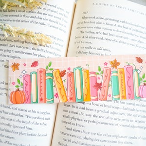 May include: A pink and peach bookmark with a stack of colorful books, a pumpkin, and fall leaves. The bookmark has a grid pattern and a sparkly design.
