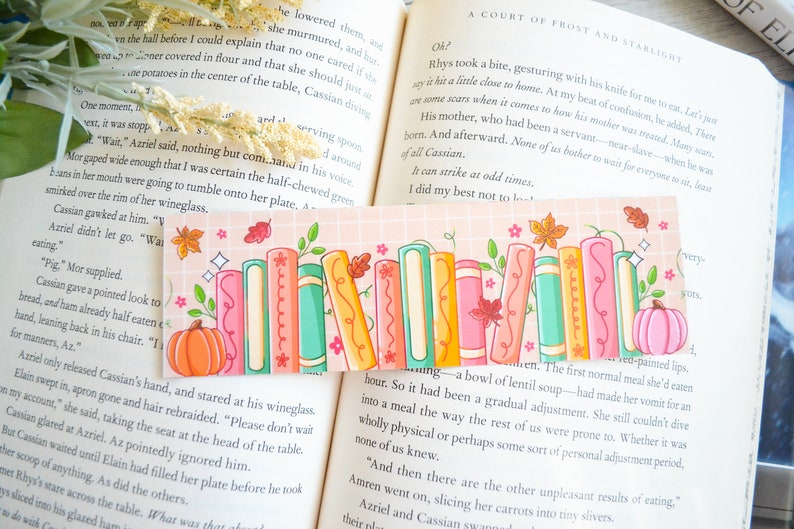 May include: A pink and peach bookmark with a stack of colorful books, a pumpkin, and fall leaves. The bookmark has a grid pattern and a sparkly design.