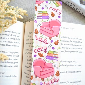 May include: A pink and white bookmark with a cozy reading theme. It features a pink armchair, stacks of books, and the words "reads cozy" in a handwritten font.