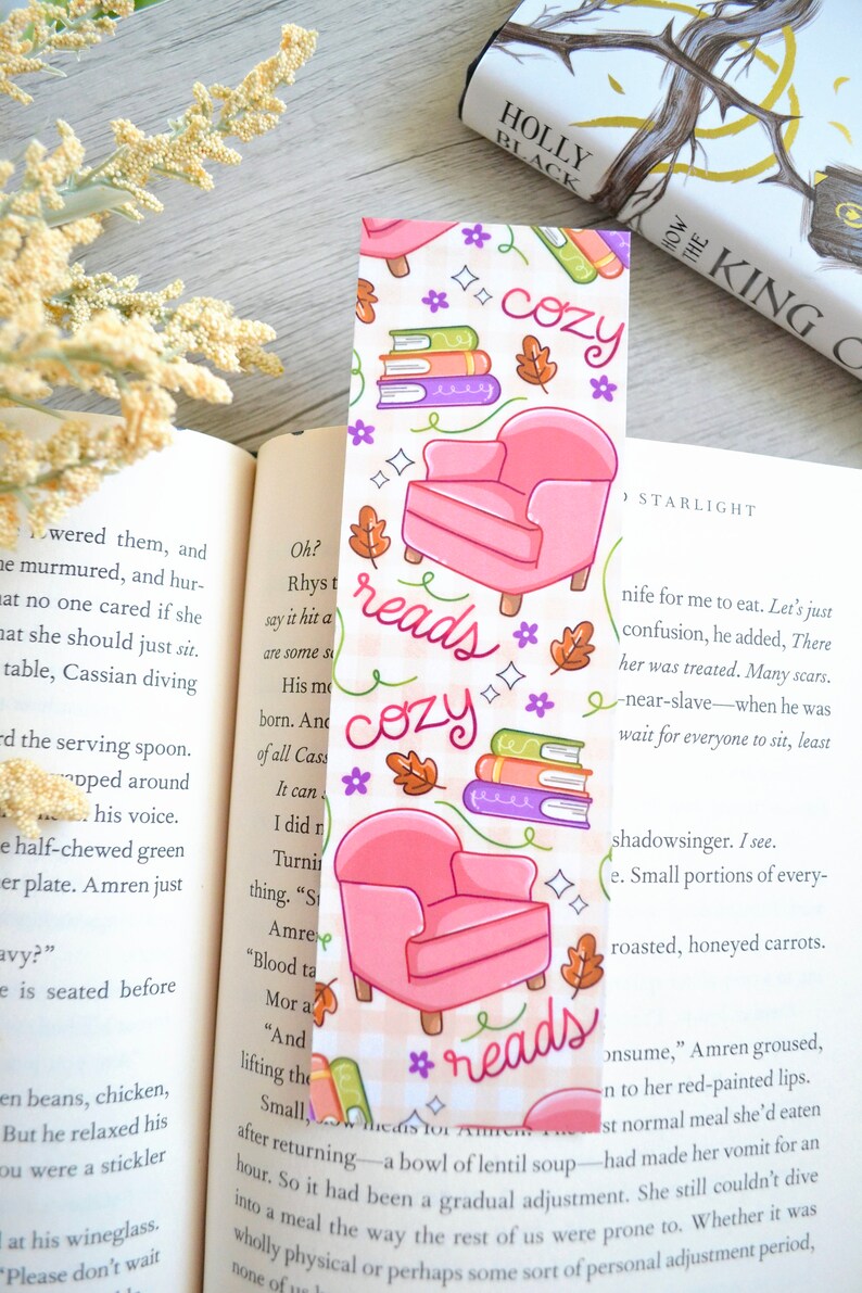 May include: A pink and white bookmark with a cozy reading theme. It features a pink armchair, stacks of books, and the words "reads cozy" in a handwritten font.