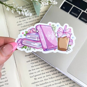 Puede incluir: Una pegatina con una pila de libros, un libro individual y un caf&eacute; helado. Los libros son rosas con detalles florales, y el caf&eacute; tiene un lazo rosa. La pegatina est&aacute; sobre un port&aacute;til.