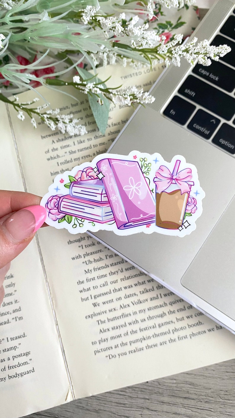 Puede incluir: Una pegatina con una pila de libros, un libro individual y un caf&eacute; helado. Los libros son rosas con detalles florales, y el caf&eacute; tiene un lazo rosa. La pegatina est&aacute; sobre un port&aacute;til.