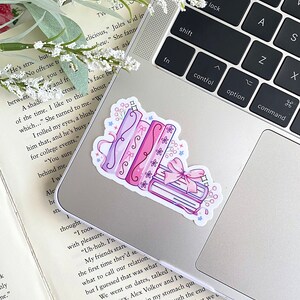 May include: A sticker featuring a stack of books and rolled scrolls in shades of pink and purple, adorned with bows and floral accents. The sticker is placed on a silver laptop.