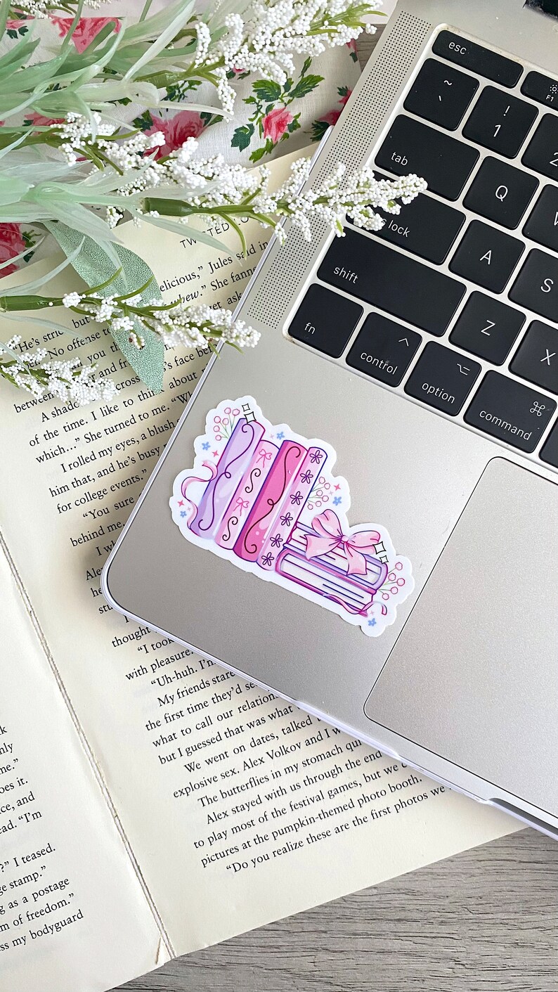 May include: A sticker featuring a stack of books and rolled scrolls in shades of pink and purple, adorned with bows and floral accents. The sticker is placed on a silver laptop.