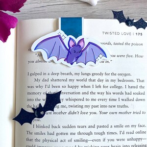 May include: A close-up of a book page with text about a person's experience with grief and loss. The text is highlighted with a purple bat bookmark with glitter accents. The book cover is pink with the word "SPOOK" in black letters.