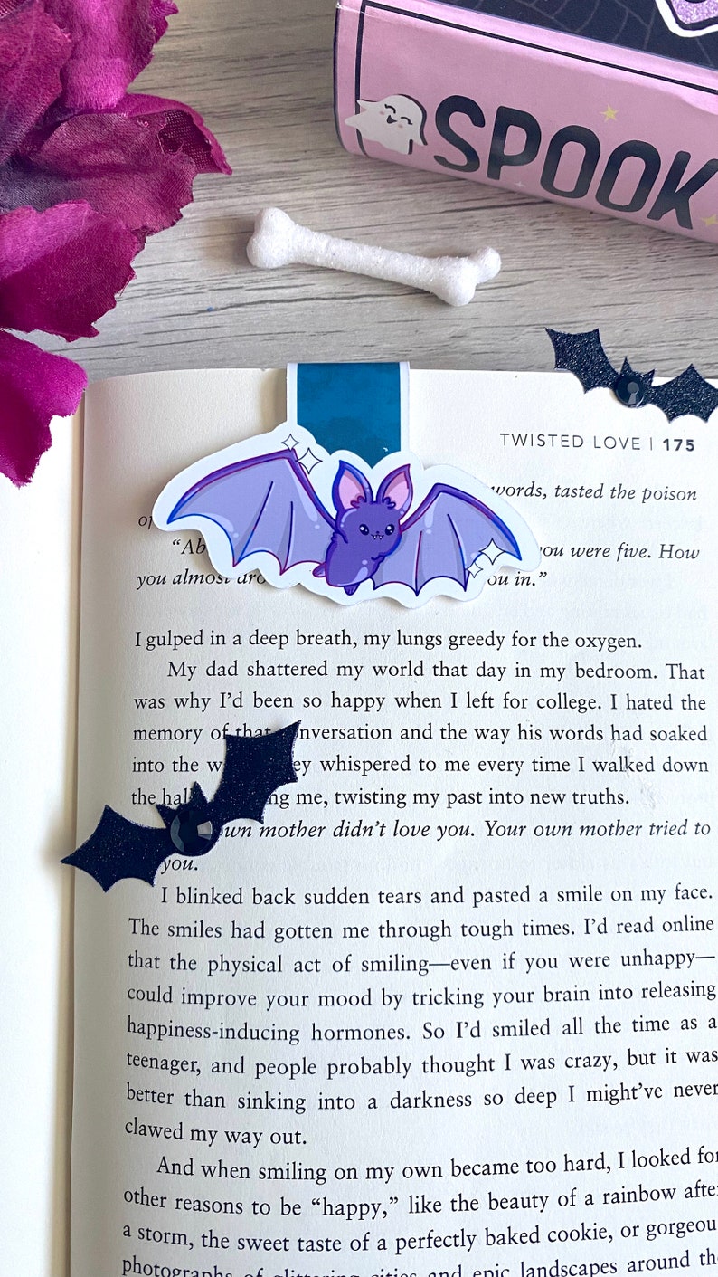 May include: A close-up of a book page with text about a person's experience with grief and loss. The text is highlighted with a purple bat bookmark with glitter accents. The book cover is pink with the word "SPOOK" in black letters.