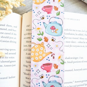 May include: A pink and white bookmark with a teacup and teapot design. The bookmark features a blue teapot with a red mushroom on top, a yellow teapot with white flowers, and a pink teacup with a brown tea inside. The bookmark also has bees, flowers, and stars.