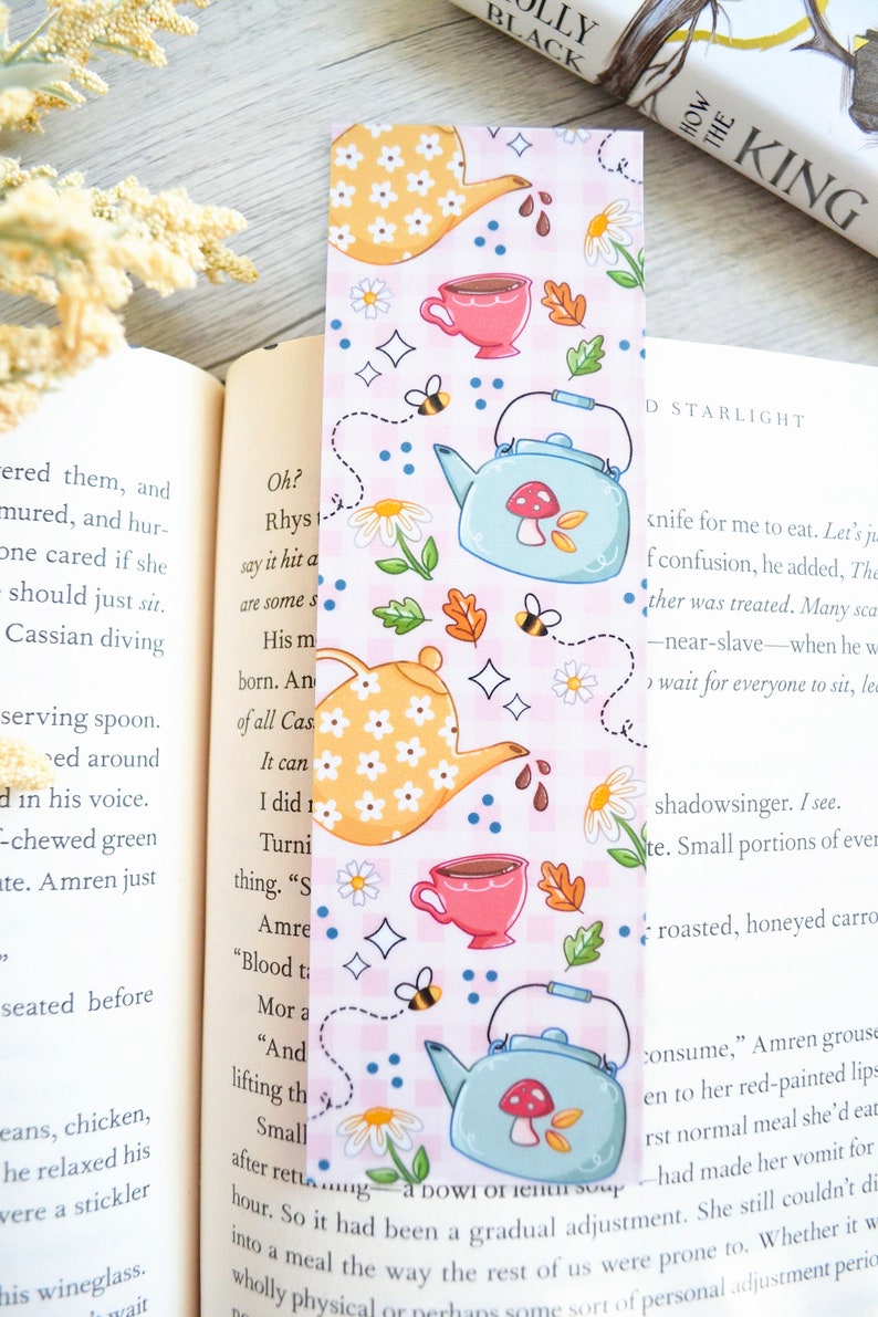 May include: A pink and white bookmark with a teacup and teapot design. The bookmark features a blue teapot with a red mushroom on top, a yellow teapot with white flowers, and a pink teacup with a brown tea inside. The bookmark also has bees, flowers, and stars.