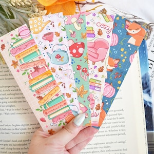 May include: Set of six colorful bookmarks featuring autumnal themes. The bookmarks feature illustrations of books, pumpkins, teacups, mushrooms, foxes, and a cozy armchair. The bookmarks are printed on a white background with a pink, blue, green, and orange color scheme.