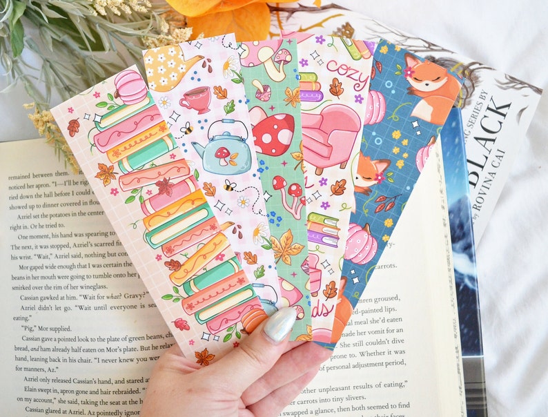May include: Set of six colorful bookmarks featuring autumnal themes. The bookmarks feature illustrations of books, pumpkins, teacups, mushrooms, foxes, and a cozy armchair. The bookmarks are printed on a white background with a pink, blue, green, and orange color scheme.