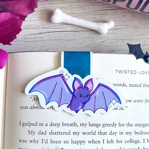 May include: A close-up of a book with a purple bat bookmark. The book has the words "Twisted Love" and "You were fine" on the page. The bookmark is a cute cartoon bat with sparkly details.