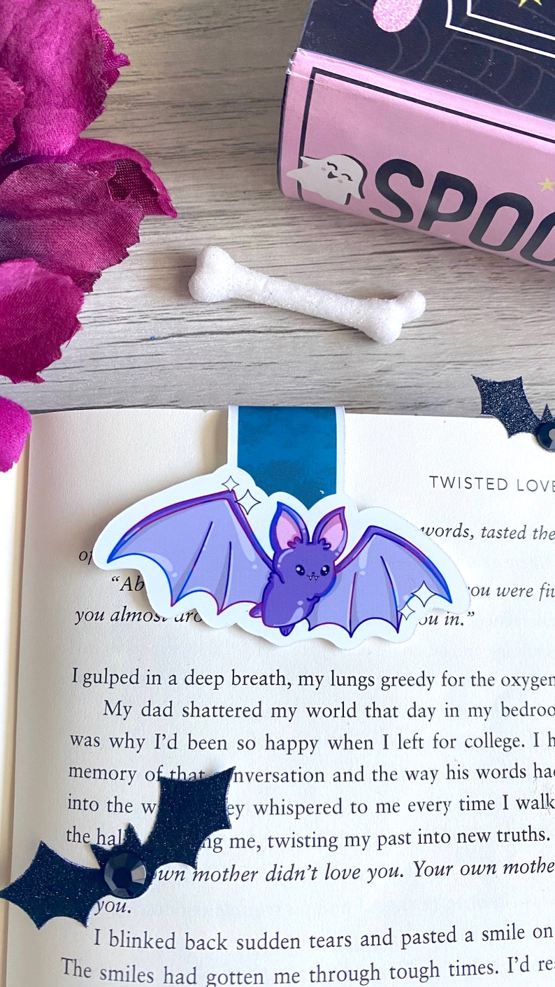 May include: A close-up of a book with a purple bat bookmark. The book has the words "Twisted Love" and "You were fine" on the page. The bookmark is a cute cartoon bat with sparkly details.