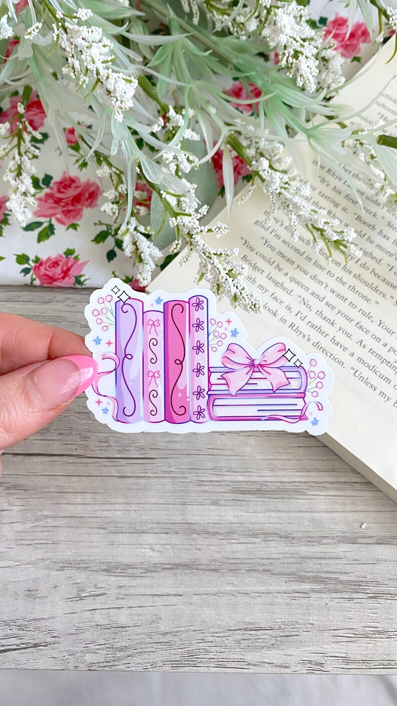May include: A sticker featuring a stack of books in shades of pink and purple, adorned with bows and floral accents. The design includes a ribbon and sparkling stars, creating a whimsical aesthetic. The sticker is set against a backdrop of a book and floral arrangement.