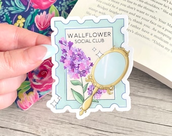 May include: A light blue sticker with a white border featuring a hand-drawn illustration of a purple flower and a gold mirror with a blue background. The sticker has the text "WALLFLOWER SOCIAL CLUB" in black text.