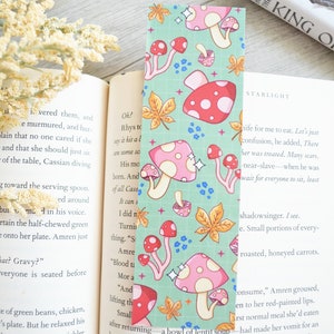 May include: A bookmark with a green background and a repeating pattern of pink and red mushrooms with white polka dots. The mushrooms have white stems and are surrounded by small blue flowers and gold leaves. The bookmark has a sparkly finish.
