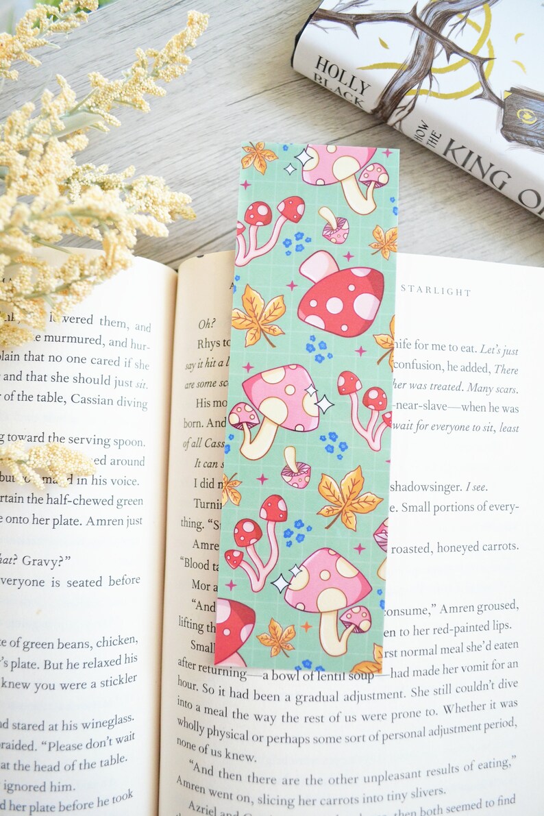 May include: A bookmark with a green background and a repeating pattern of pink and red mushrooms with white polka dots. The mushrooms have white stems and are surrounded by small blue flowers and gold leaves. The bookmark has a sparkly finish.