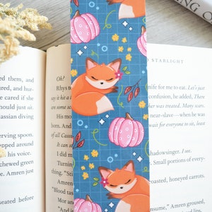 May include: A blue bookmark with a repeating pattern of orange foxes sleeping on pink pumpkins. The background is a blue grid with white stars and yellow flowers.