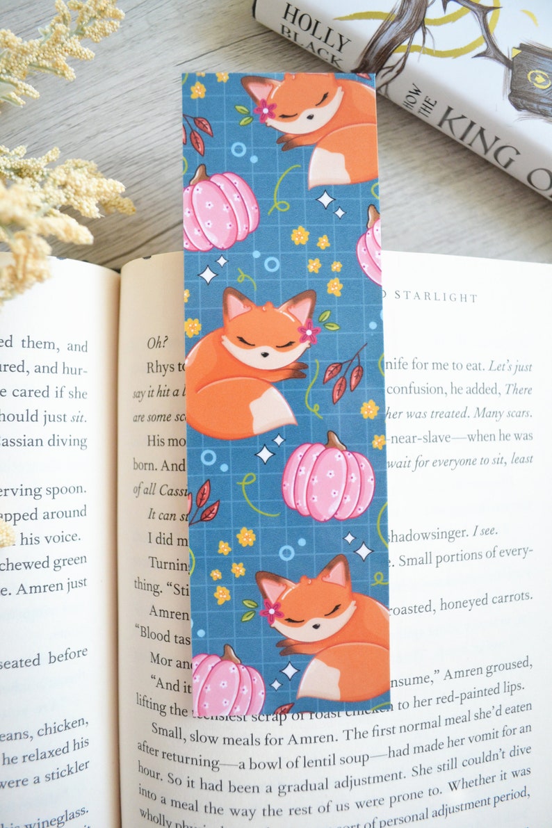 May include: A blue bookmark with a repeating pattern of orange foxes sleeping on pink pumpkins. The background is a blue grid with white stars and yellow flowers.