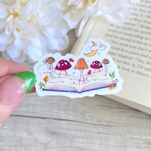 May include: A sticker featuring an open book with a crescent moon and clouds above it.  Four mushrooms with red and orange caps are growing out of the book.  The book has a rainbow-colored spine and purple pages.