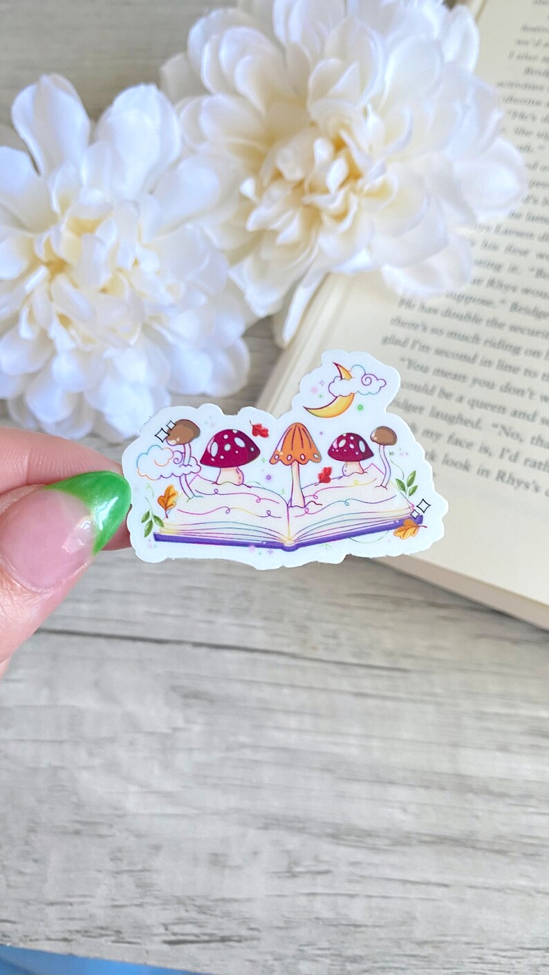 May include: A sticker featuring an open book with a crescent moon and clouds above it.  Four mushrooms with red and orange caps are growing out of the book.  The book has a rainbow-colored spine and purple pages.