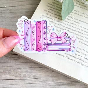May include: A sticker featuring a stack of books and several upright books in shades of pink and purple. The books are decorated with bows, flowers, and decorative lines. The sticker is on a book page.