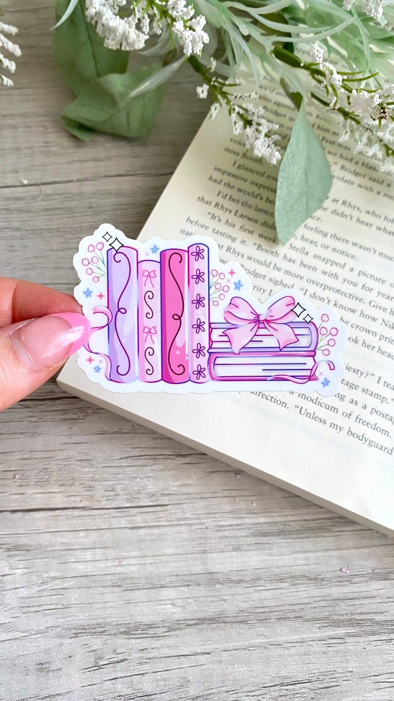 May include: A sticker featuring a stack of books and several upright books in shades of pink and purple. The books are decorated with bows, flowers, and decorative lines. The sticker is on a book page.