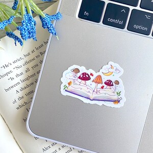May include: A sticker of an open book with three mushrooms growing out of it. The book is purple and the mushrooms are red and orange. There are leaves and a crescent moon in the background.