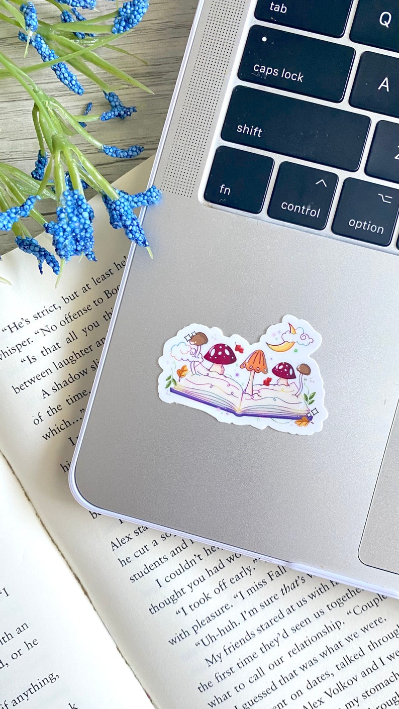 May include: A sticker of an open book with three mushrooms growing out of it. The book is purple and the mushrooms are red and orange. There are leaves and a crescent moon in the background.