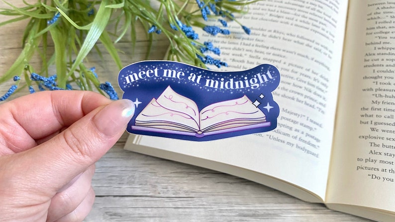 May include: A blue and white sticker with a book and stars. The text on the sticker reads "meet me at midnight".