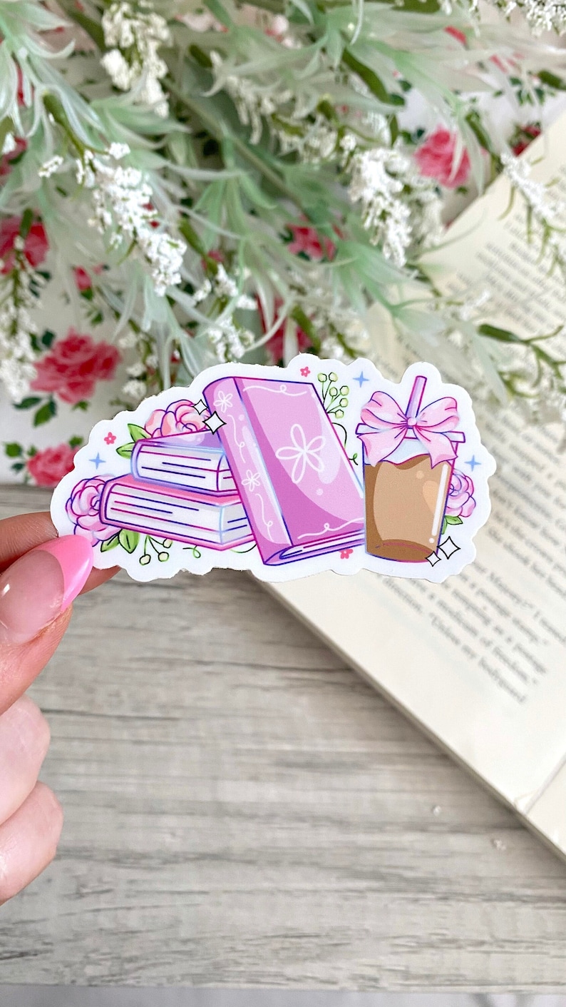 Puede incluir: Una pegatina con una pila de libros rosas, una flor rosa y una bebida de caf&eacute; con un lazo rosa. La pegatina est&aacute; rodeada de flores rosas y hojas verdes. La pegatina est&aacute; sujeta por una persona.