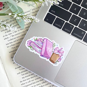 Puede incluir: Una pegatina con una pila de libros, un diario y una bebida de caf&eacute; con un lazo rosa. Los libros y el diario son morados con detalles florales. La pegatina est&aacute; colocada en un ordenador port&aacute;til.