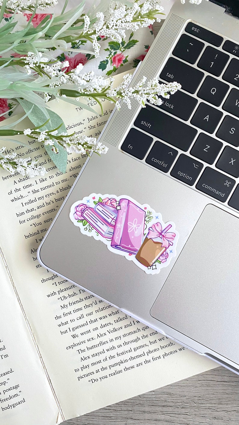 Puede incluir: Una pegatina con una pila de libros, un diario y una bebida de caf&eacute; con un lazo rosa. Los libros y el diario son morados con detalles florales. La pegatina est&aacute; colocada en un ordenador port&aacute;til.