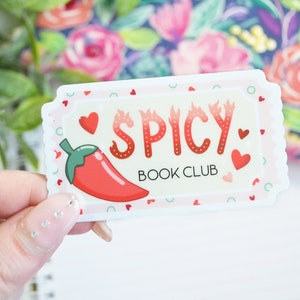 May include: A white sticker with a red chili pepper and the text "Spicy Book Club" in red letters with flames. The sticker is decorated with small red hearts and white polka dots.